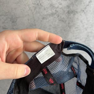 May include: A blue denim trucker hat with a mesh back panel. A white interior tag displays the text: 65% POLYESTER, 35% COTTON, MADE IN CHINA, ONE SIZE FITS ALL. The inner black sweatband features repeating red text branding throughout the interior.