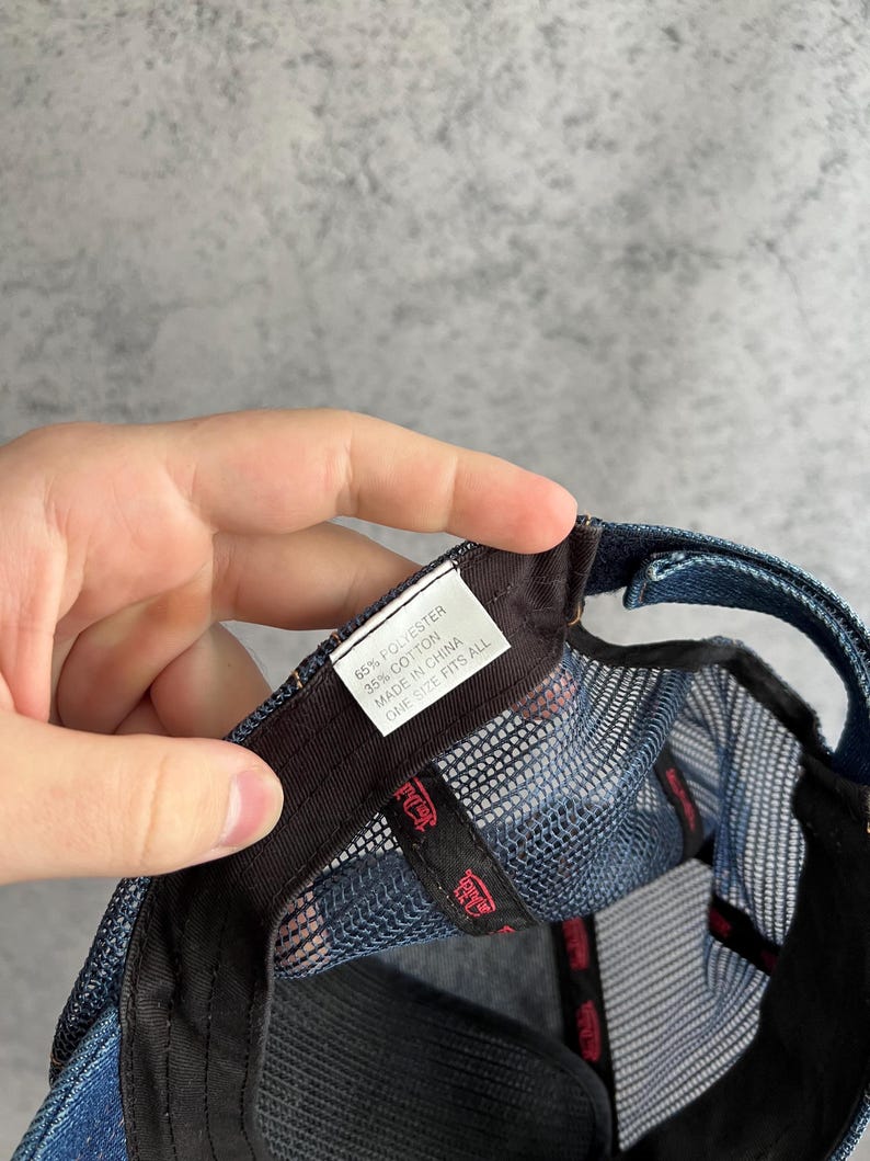 May include: A blue denim trucker hat with a mesh back panel. A white interior tag displays the text: 65% POLYESTER, 35% COTTON, MADE IN CHINA, ONE SIZE FITS ALL. The inner black sweatband features repeating red text branding throughout the interior.