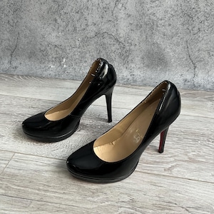 May include: A pair of black patent leather pumps with a rounded toe, a high stiletto heel, and a beige interior lining. The shoes feature a small platform under the toe area and a distinct red accent on the bottom of the heel tip.
