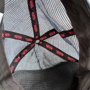 May include: The interior of a blue denim trucker hat featuring a dark blue mesh back panel. Black fabric tape lines the inner seams, printed with the red Von Dutch logo repeatedly. A black sweatband lines the bottom edge, and a metal button sits at the crown.
