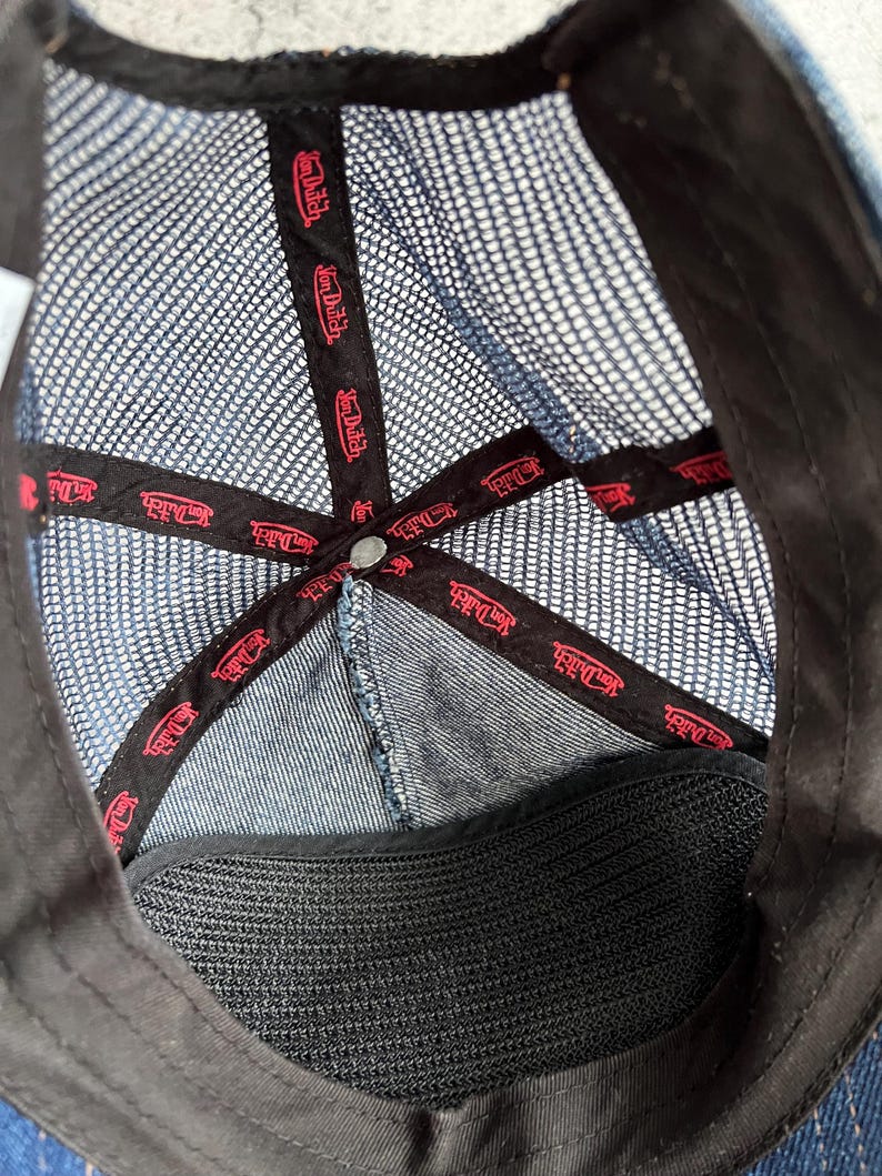 May include: The interior of a blue denim trucker hat featuring a dark blue mesh back panel. Black fabric tape lines the inner seams, printed with the red Von Dutch logo repeatedly. A black sweatband lines the bottom edge, and a metal button sits at the crown.