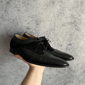 Pode incluir: Um par de sapatos de couro preto com um painel de camurça na parte superior. Os sapatos têm um design clássico com bico fino e fecho de atacadores. Os sapatos são segurados por uma mão.