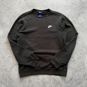 May include: Dark gray crew neck sweatshirt with the Nike logo in white on the chest. The sweatshirt has long sleeves and a small tag near the collar. The fabric appears to be a soft, comfortable material.