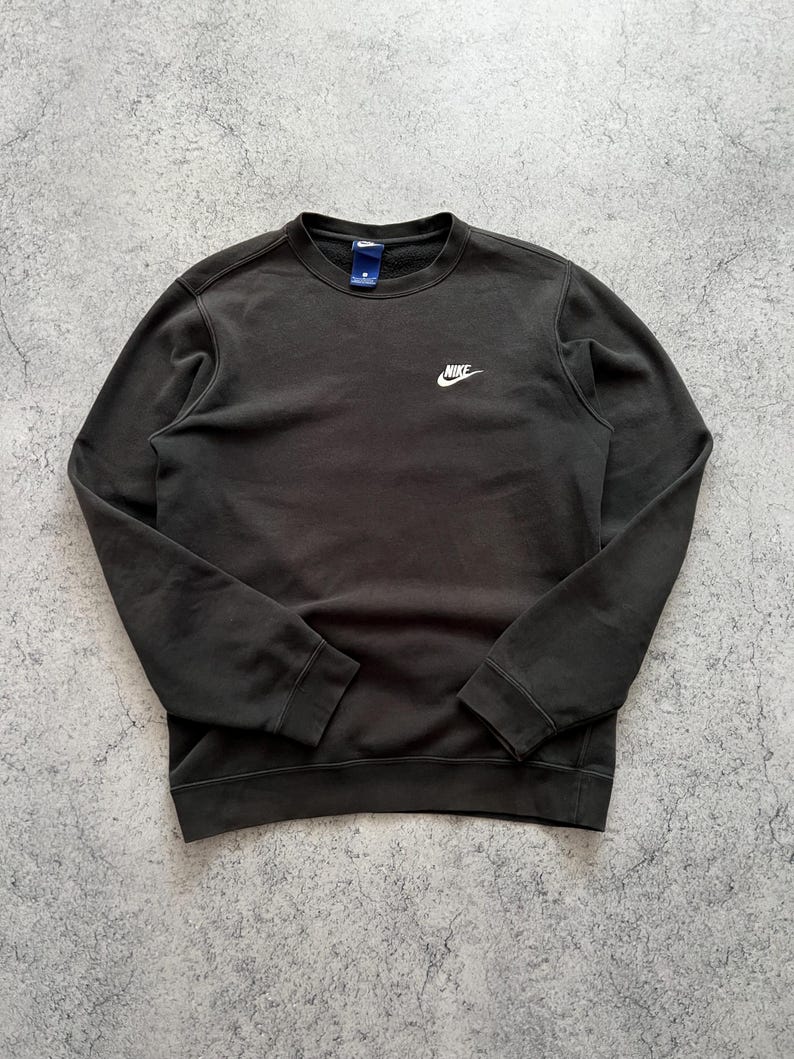 May include: Dark gray crew neck sweatshirt with the Nike logo in white on the chest. The sweatshirt has long sleeves and a small tag near the collar. The fabric appears to be a soft, comfortable material.