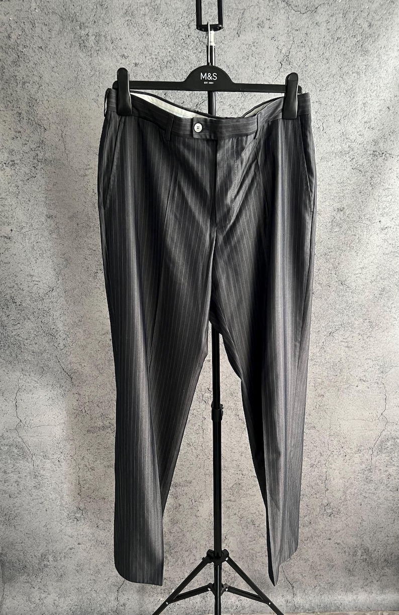 May include: Dark gray trousers featuring a subtle vertical pinstripe pattern, a button closure at the waistband, and side pockets. The trousers are displayed on a black M&S branded hanger, showcasing a straight-leg cut and a tailored appearance.