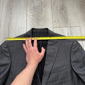 May include: A charcoal gray blazer with thin white pinstripes, featuring a notched lapel and a black interior lining. A yellow measuring tape is positioned across the shoulders, indicating a width of approximately 20 inches from edge to edge.