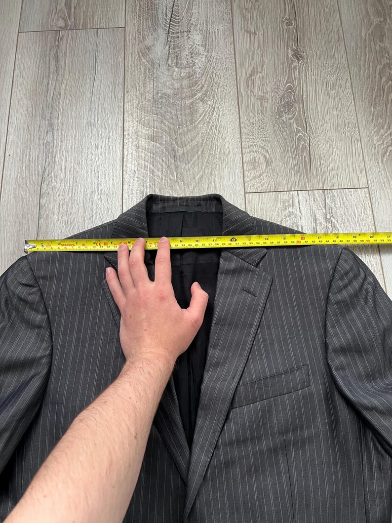 May include: A charcoal gray blazer with thin white pinstripes, featuring a notched lapel and a black interior lining. A yellow measuring tape is positioned across the shoulders, indicating a width of approximately 20 inches from edge to edge.