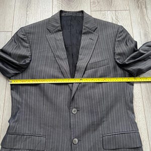 May include: A charcoal gray pinstriped blazer with a black interior lining featuring a repeating logo pattern. The blazer has a notched lapel, two front buttons, and two flap pockets. A yellow measuring tape shows a width of approximately 23 inches.