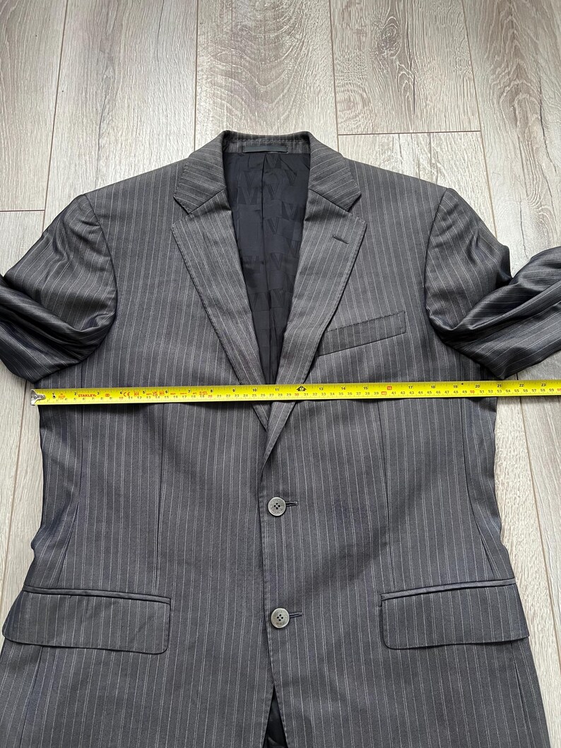 May include: A charcoal gray pinstriped blazer with a black interior lining featuring a repeating logo pattern. The blazer has a notched lapel, two front buttons, and two flap pockets. A yellow measuring tape shows a width of approximately 23 inches.