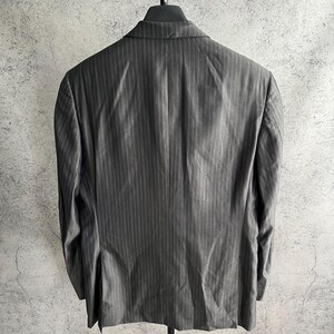 May include: A charcoal gray blazer featuring a subtle vertical pinstripe pattern. The garment is displayed from the back, showing a structured collar, long sleeves, and a single vent at the bottom hem, hanging on a black hanger against a textured wall.