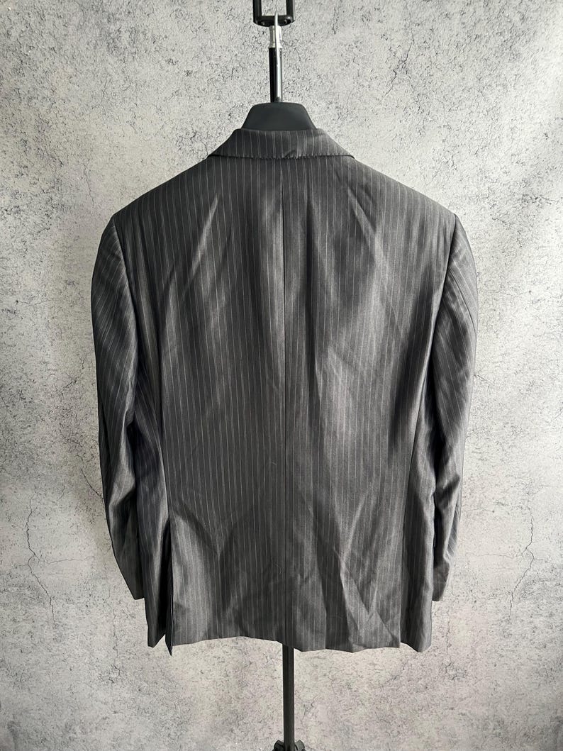 May include: A charcoal gray blazer featuring a subtle vertical pinstripe pattern. The garment is displayed from the back, showing a structured collar, long sleeves, and a single vent at the bottom hem, hanging on a black hanger against a textured wall.