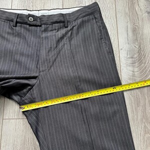May include: Dark gray trousers with thin white pinstripes, featuring a button closure and a waistband. A yellow measuring tape is placed across the front, showing a width measurement of approximately 16 inches across the thigh area.