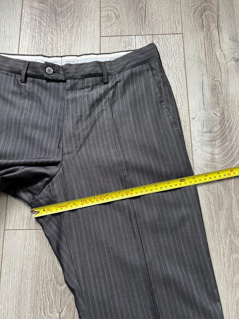 May include: Dark gray trousers with thin white pinstripes, featuring a button closure and a waistband. A yellow measuring tape is placed across the front, showing a width measurement of approximately 16 inches across the thigh area.