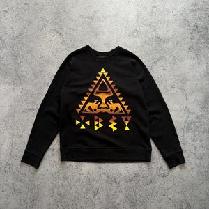 Może przedstawiać: Czarna bluza z okrągłym dekoltem z geometrycznym wzorem w kolorach pomarańczowym, żółtym i brązowym. Wzór ma kształt trójkąta z obrazem przypominającym twarz pośrodku. Bluza ma długie rękawy i okrągły dekolt.