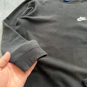 May include: A dark gray Nike sweatshirt with a small white Nike logo on the chest. The sweatshirt has a crew neck and ribbed cuffs. The fabric appears to be slightly faded, giving it a vintage look.