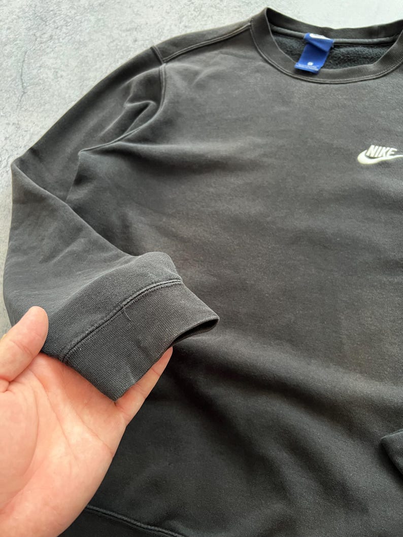 May include: A dark gray Nike sweatshirt with a small white Nike logo on the chest. The sweatshirt has a crew neck and ribbed cuffs. The fabric appears to be slightly faded, giving it a vintage look.