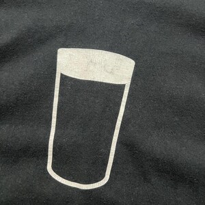 May include: Black t-shirt featuring a white outline of a pint glass. The graphic is centered on the chest. The shirt is made of a soft, dark fabric. The design is simple and bold, suitable for casual wear.