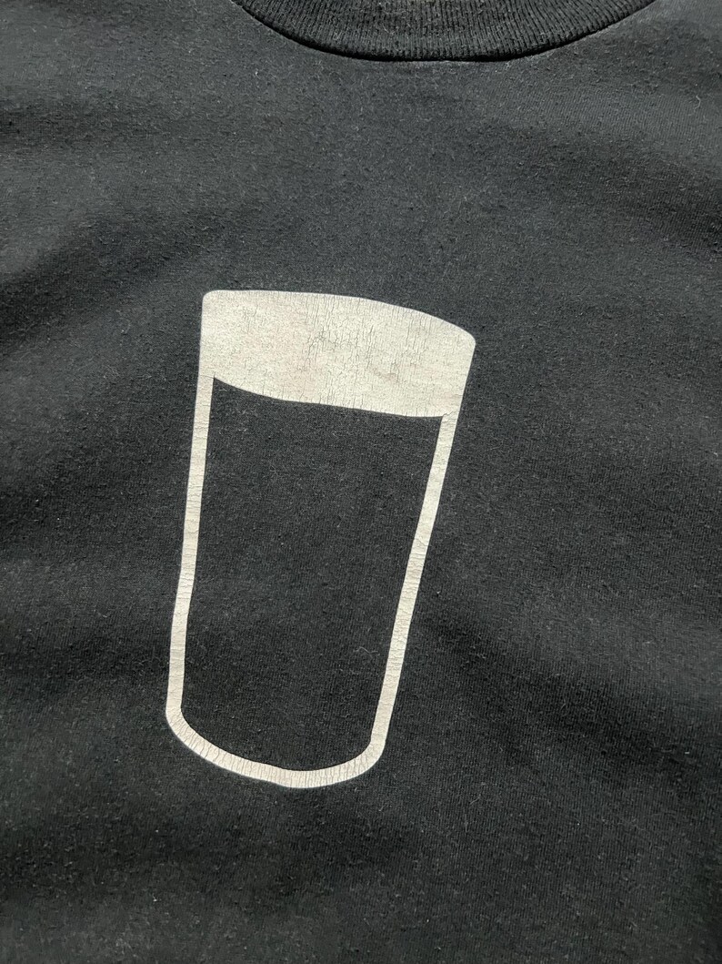 May include: Black t-shirt featuring a white outline of a pint glass. The graphic is centered on the chest. The shirt is made of a soft, dark fabric. The design is simple and bold, suitable for casual wear.