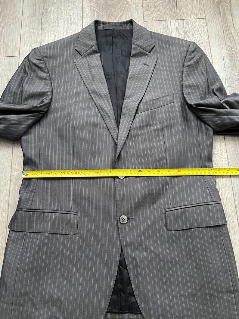 May include: A grey pinstriped blazer with a black interior lining, featuring two front buttons and two flap pockets. A yellow measuring tape is placed across the midsection, indicating a width of approximately 22 inches across the chest.