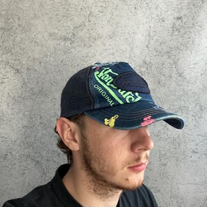 May include: A dark blue denim baseball cap with a mesh back, featuring neon green text that reads Von Dutch Original, a dark blue rectangular patch, and small colorful graphic icons printed across the front panel and the curved brim.