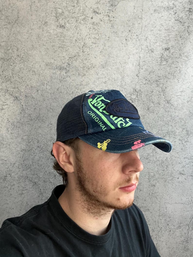 May include: A dark blue denim baseball cap with a mesh back, featuring neon green text that reads Von Dutch Original, a dark blue rectangular patch, and small colorful graphic icons printed across the front panel and the curved brim.