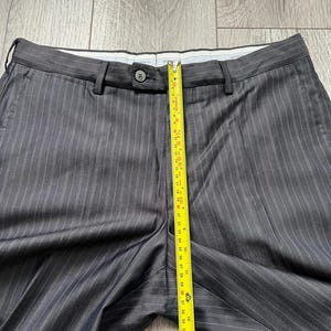May include: Dark gray trousers with thin white pinstripes, featuring a button closure at the waistband and belt loops. A yellow measuring tape is placed vertically over the center, showing a length measurement of approximately 15 inches.