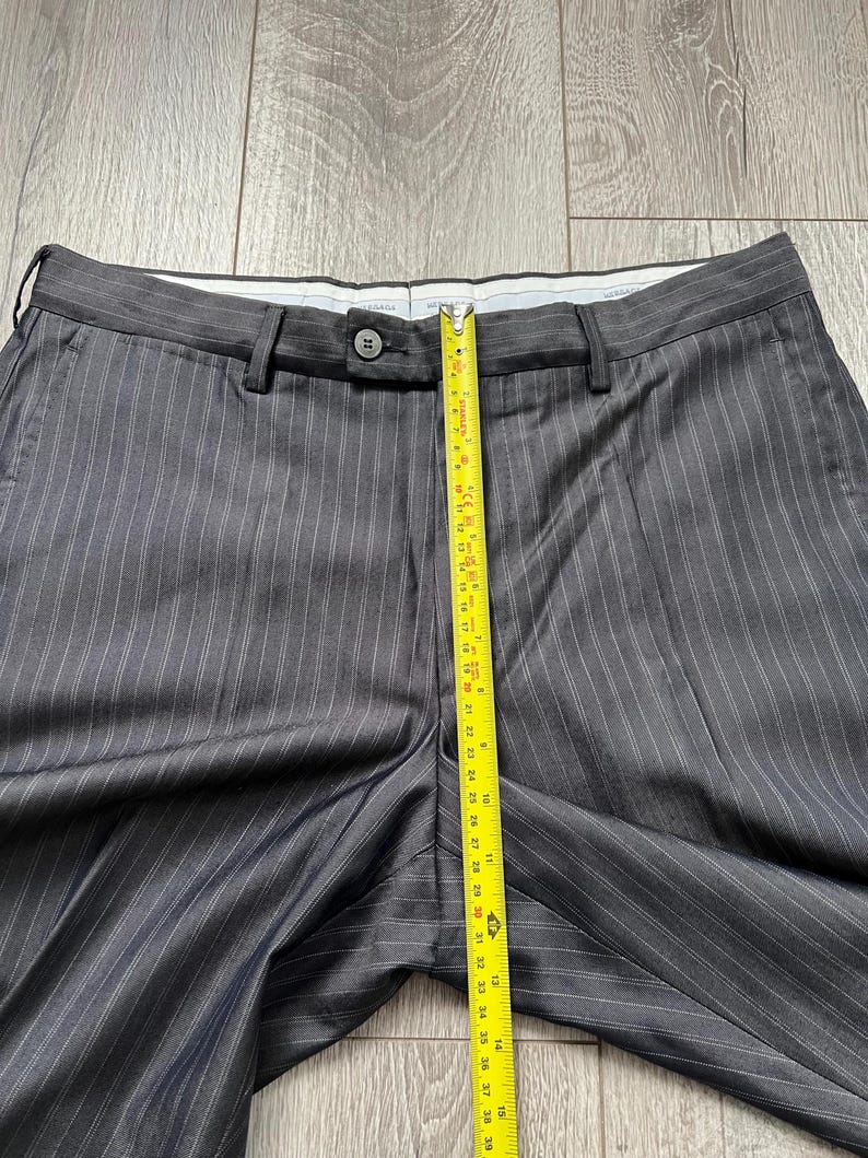 May include: Dark gray trousers with thin white pinstripes, featuring a button closure at the waistband and belt loops. A yellow measuring tape is placed vertically over the center, showing a length measurement of approximately 15 inches.