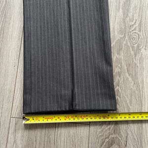 May include: A pair of dark gray trousers featuring a subtle white pinstripe pattern. The bottom hem of the pant leg is positioned against a yellow measuring tape, which indicates a width of approximately 9.5 inches across the flat fabric.