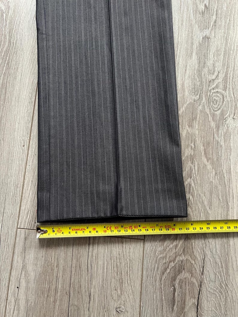 May include: A pair of dark gray trousers featuring a subtle white pinstripe pattern. The bottom hem of the pant leg is positioned against a yellow measuring tape, which indicates a width of approximately 9.5 inches across the flat fabric.