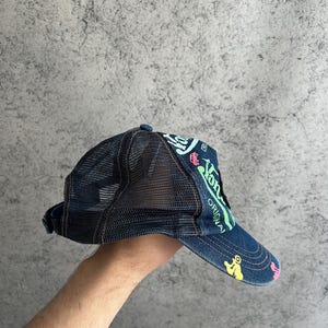 May include: A dark blue denim trucker hat featuring a black mesh back panel and a curved brim. The front and brim are decorated with colorful graphic prints in neon green, light blue, yellow, and pink, including the text "Von Dutch" and "ORIGINAL".