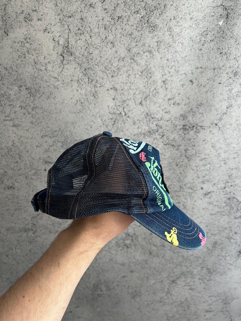 May include: A dark blue denim trucker hat featuring a black mesh back panel and a curved brim. The front and brim are decorated with colorful graphic prints in neon green, light blue, yellow, and pink, including the text "Von Dutch" and "ORIGINAL".