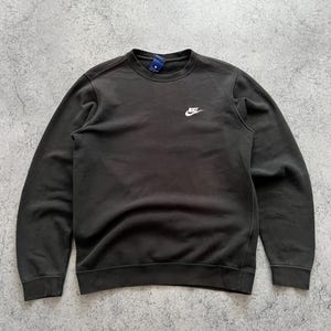 May include: Dark gray crew neck sweatshirt with the Nike logo in white on the upper left chest. The sweatshirt has a simple, classic design, with ribbed cuffs and hem. A small tag is visible at the back of the neck.
