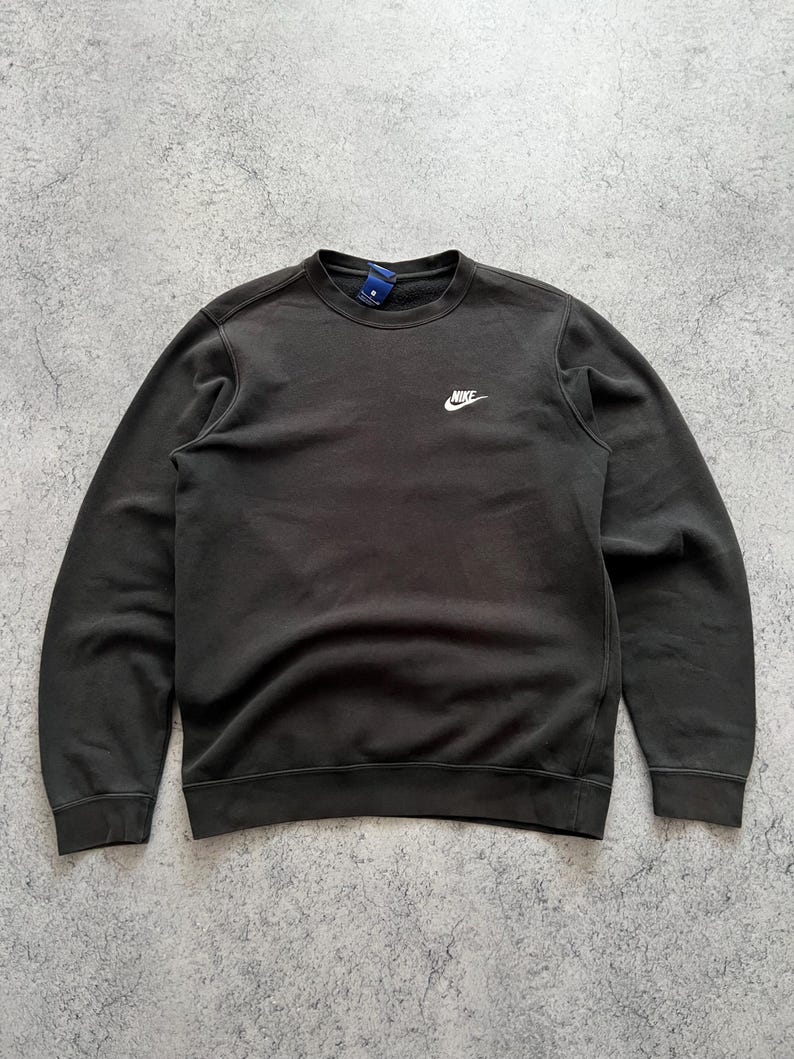 May include: Dark gray crew neck sweatshirt with the Nike logo in white on the upper left chest. The sweatshirt has a simple, classic design, with ribbed cuffs and hem. A small tag is visible at the back of the neck.