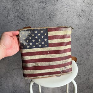 May include: A rectangular crossbody bag with a design of the American flag. The bag has a distressed appearance with red and white stripes and a blue field with white stars. It has a zip closure and a brown strap.