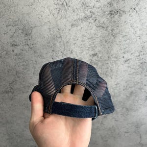 May include: A dark blue denim trucker hat featuring a mesh back panel and an adjustable strap closure. The cap is constructed with visible brown stitching along the seams and a structured crown, showcasing a classic design with breathable mesh.