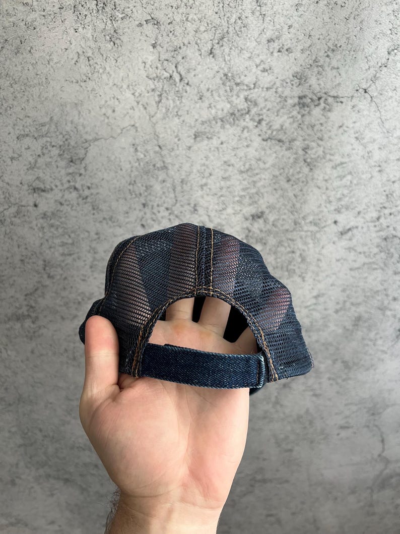 May include: A dark blue denim trucker hat featuring a mesh back panel and an adjustable strap closure. The cap is constructed with visible brown stitching along the seams and a structured crown, showcasing a classic design with breathable mesh.