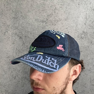 May include: A denim trucker hat featuring a dark blue mesh back and a front panel adorned with embroidered Von Dutch logos and colorful graphics of small cars and motorcycles. The brim displays the text Von Dutch Originals in white lettering.