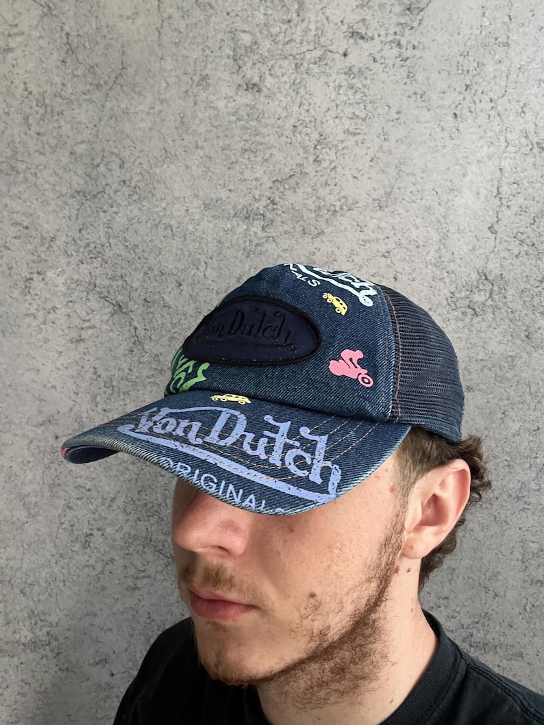 May include: A denim trucker hat featuring a dark blue mesh back and a front panel adorned with embroidered Von Dutch logos and colorful graphics of small cars and motorcycles. The brim displays the text Von Dutch Originals in white lettering.