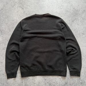 May include: A dark gray sweatshirt with a crew neck and long sleeves. The garment is made of a soft material and has ribbed cuffs and hem. The sweatshirt is displayed on a gray, textured surface.