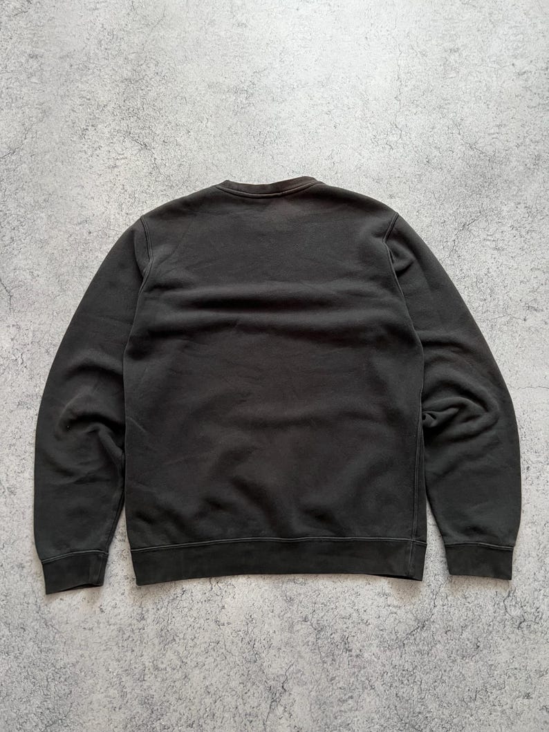 May include: A dark gray sweatshirt with a crew neck and long sleeves. The garment is made of a soft material and has ribbed cuffs and hem. The sweatshirt is displayed on a gray, textured surface.