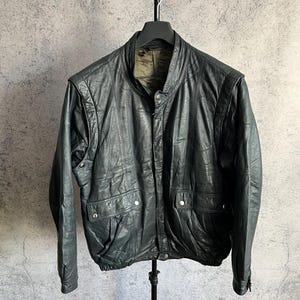 May include: Black leather jacket with a stand-up collar, two front pockets, and a button-up closure. The jacket is hanging on a black hanger against a gray background. The jacket has a ribbed hem and cuffs.