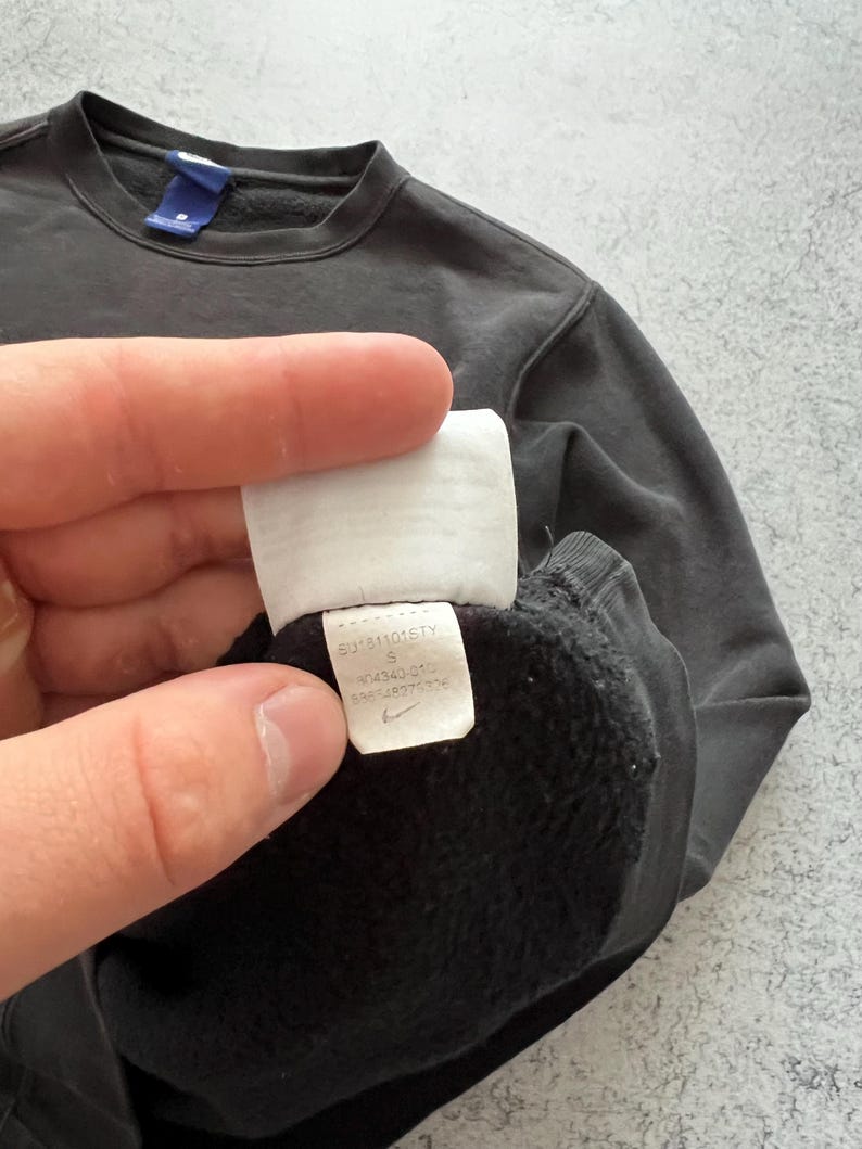 May include: Dark gray sweatshirt with a crew neck and a small blue tag. A white tag with black text and a Nike logo is visible. The sweatshirt is a size small.