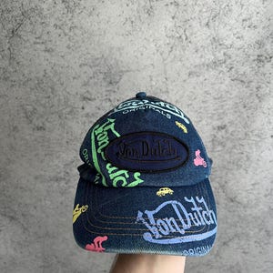 May include: A dark blue denim baseball cap featuring a central oval patch with the text Von Dutch in dark blue embroidery. The cap is decorated with colorful screen-printed graphics including the text Von Dutch and small car icons in neon green, pink, yellow, and light blue.