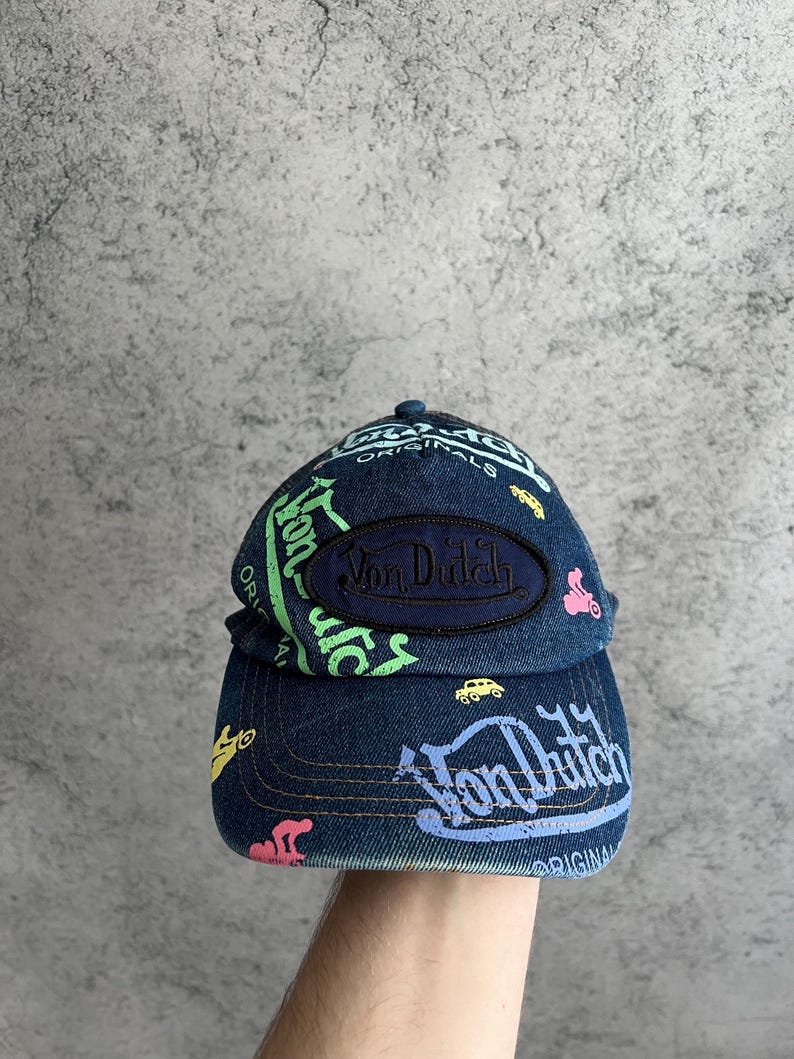 May include: A dark blue denim baseball cap featuring a central oval patch with the text Von Dutch in dark blue embroidery. The cap is decorated with colorful screen-printed graphics including the text Von Dutch and small car icons in neon green, pink, yellow, and light blue.