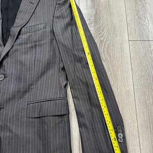 May include: A charcoal gray pinstriped blazer with a black interior lining, featuring a notched lapel, a single button closure, and two flap pockets. A yellow measuring tape placed along the sleeve shows a length of approximately 26 inches.