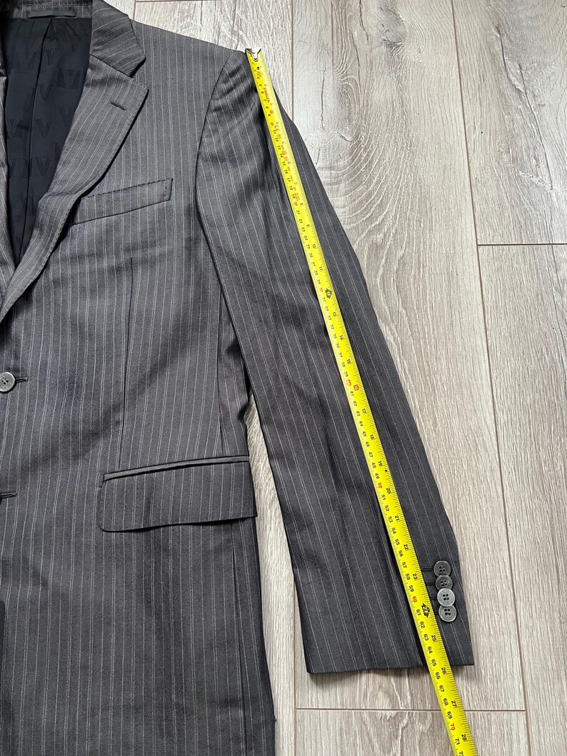 May include: A charcoal gray pinstriped blazer with a black interior lining, featuring a notched lapel, a single button closure, and two flap pockets. A yellow measuring tape placed along the sleeve shows a length of approximately 26 inches.