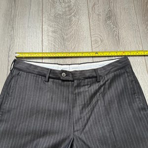May include: Dark gray trousers with thin white pinstripes, featuring a button closure, belt loops, and a white inner waistband. A yellow measuring tape placed across the top shows a waist width of approximately 18 inches from side to side.