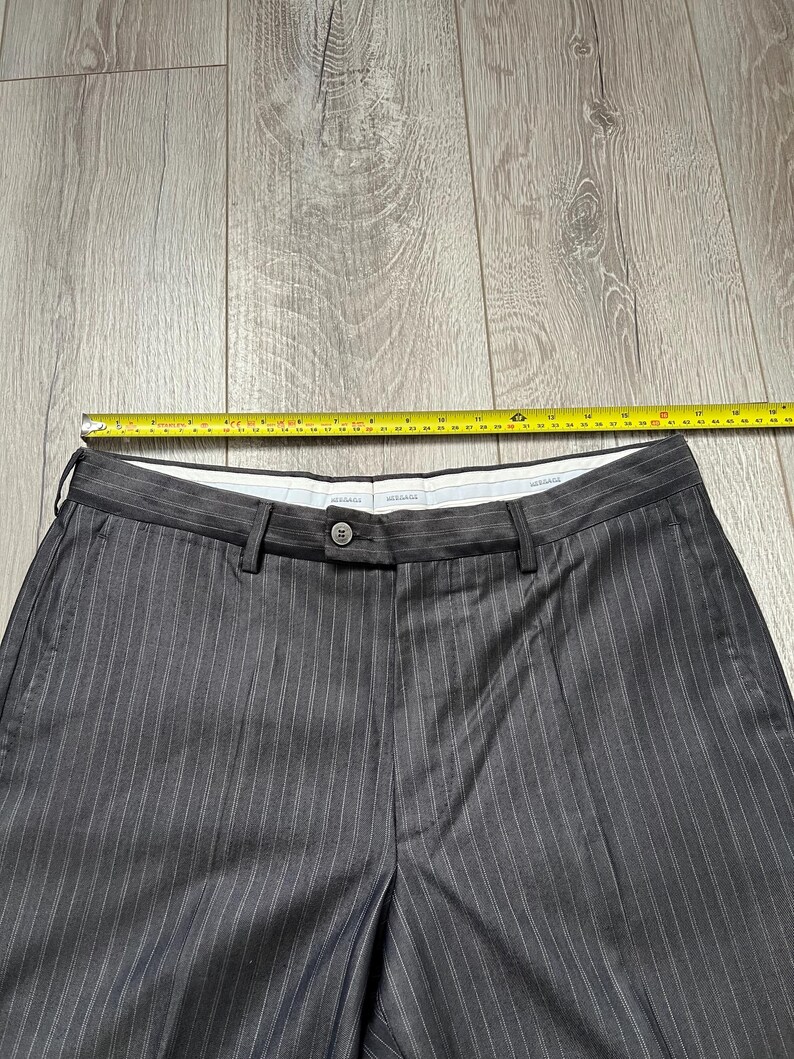 May include: Dark gray trousers with thin white pinstripes, featuring a button closure, belt loops, and a white inner waistband. A yellow measuring tape placed across the top shows a waist width of approximately 18 inches from side to side.