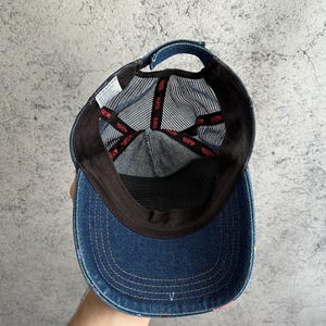 May include: A blue denim baseball cap with a mesh back panel. The interior features black fabric lining along the brim and crown, with black mesh panels. Red branded tape lines the interior seams, and a white care label is attached to the side.