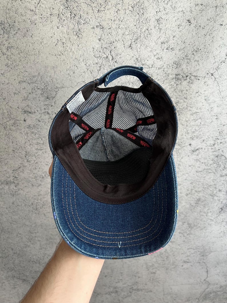 May include: A blue denim baseball cap with a mesh back panel. The interior features black fabric lining along the brim and crown, with black mesh panels. Red branded tape lines the interior seams, and a white care label is attached to the side.